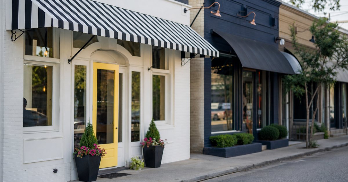 Two neighboring small shops on a quiet main street in the morning, one freshly painted with a new striped awning and flowers by the door