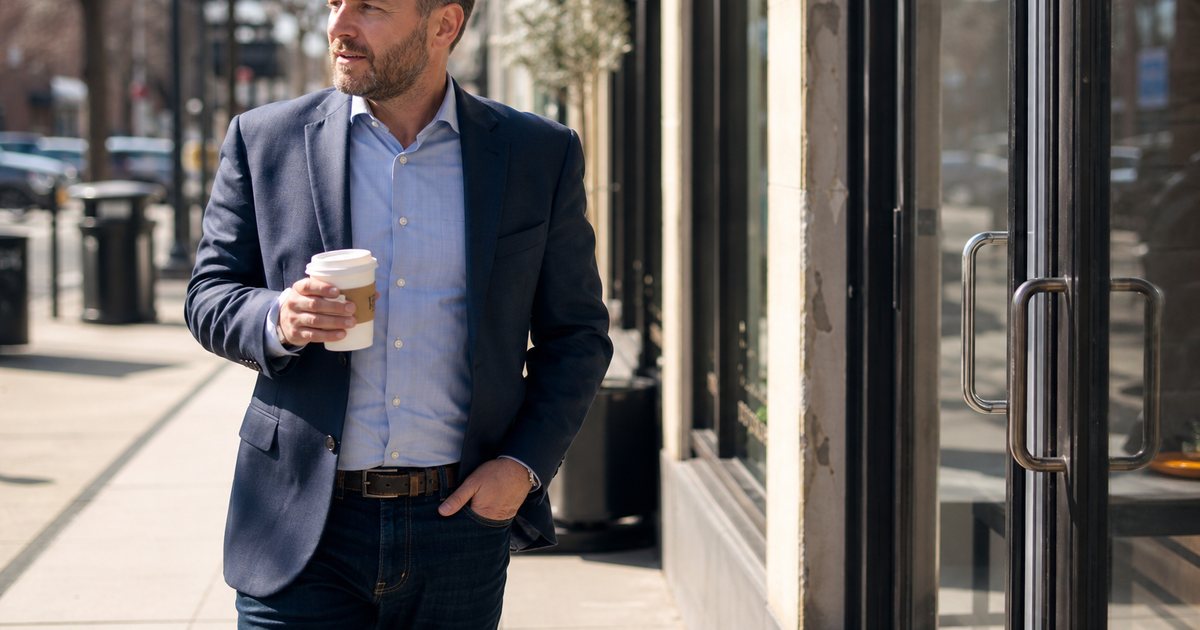 A regional manager walking between two storefront doors on a busy commercial street, coffee in hand, sunlight on the pavement